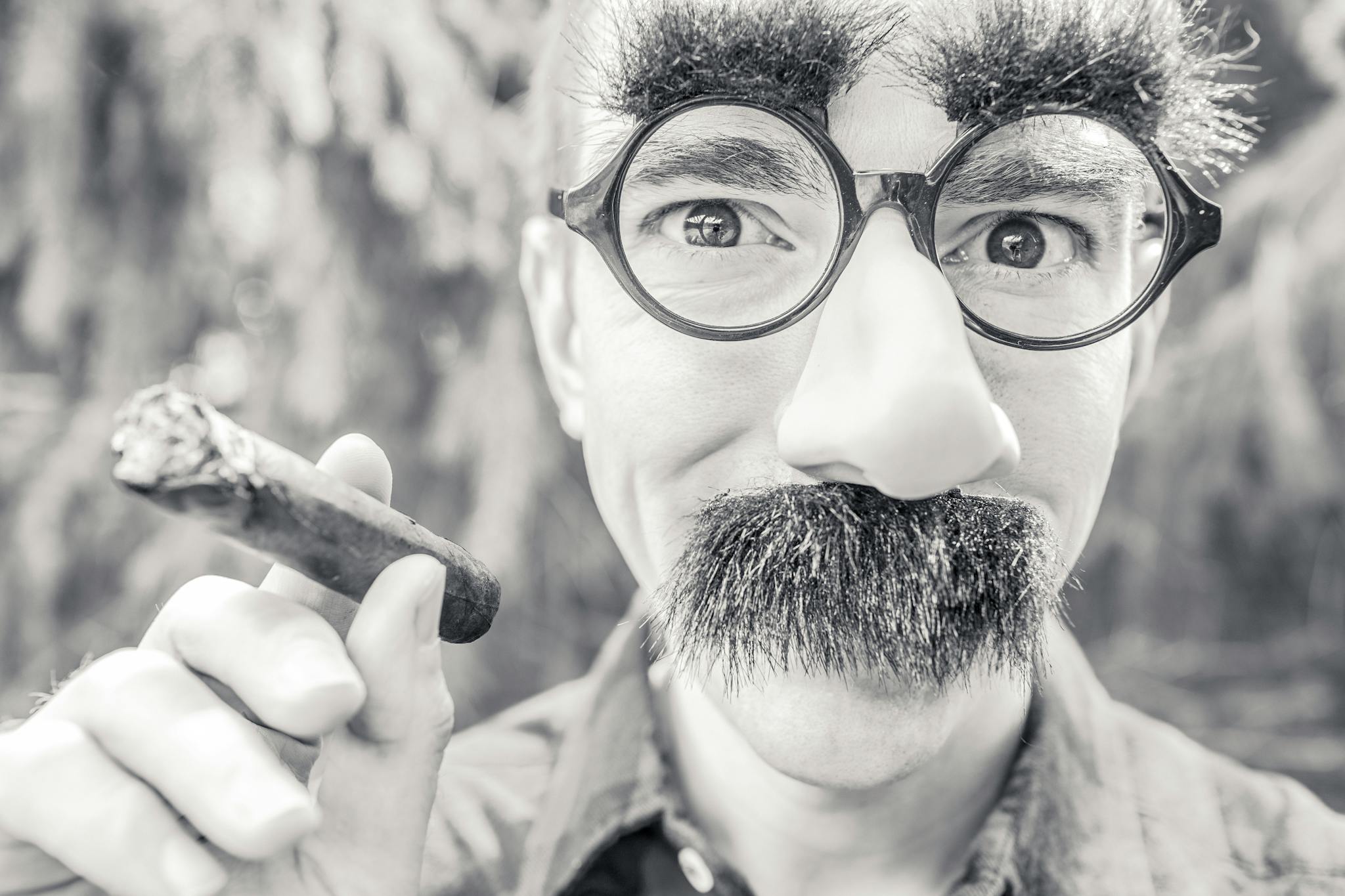 Silly portrait of a man wearing costume glasses and mustache, holding a cigar.