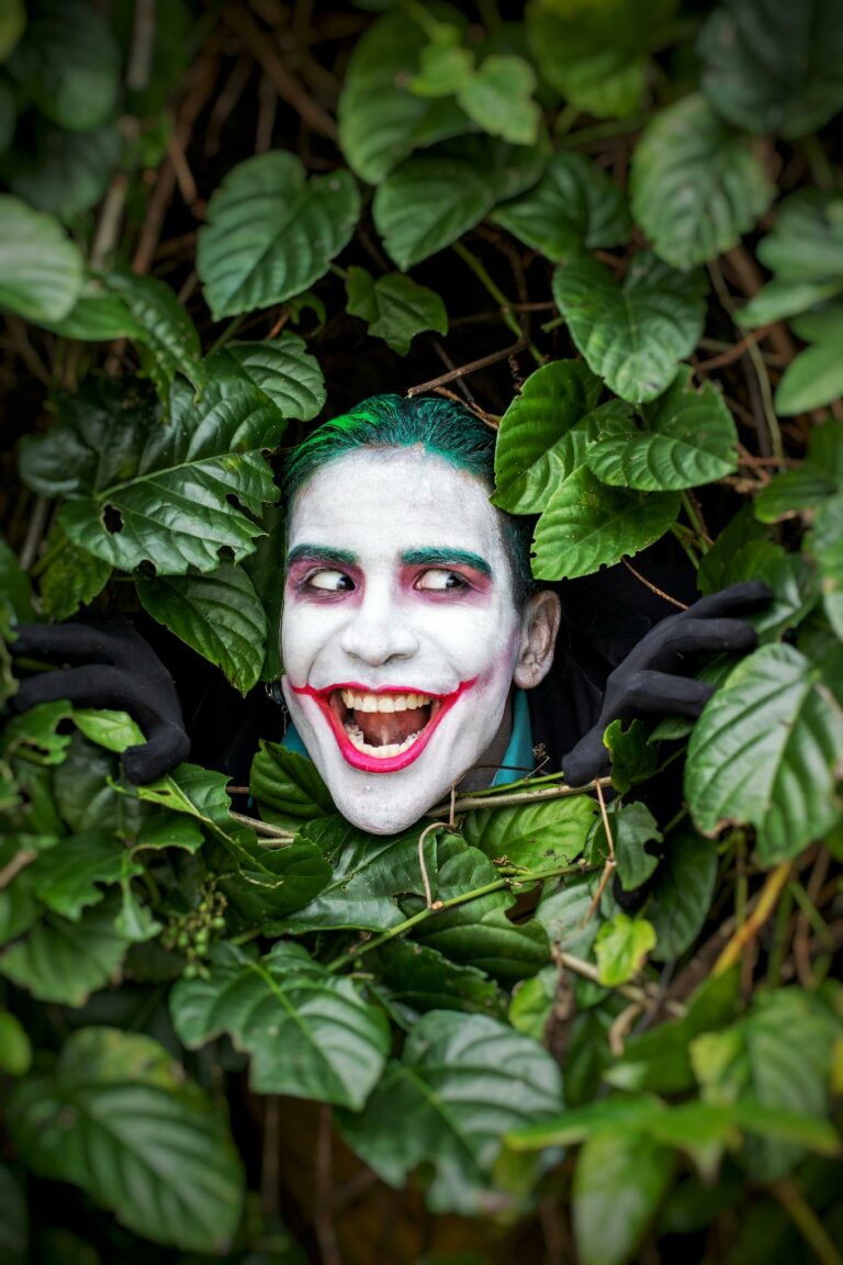 Smiling face painted like Joker amidst lush green leaves, outdoors.